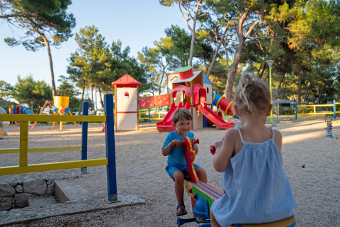 Dos niños juegan en un balancín en un colorido parque infantil con árboles en Camping Village Poljana, Croacia.