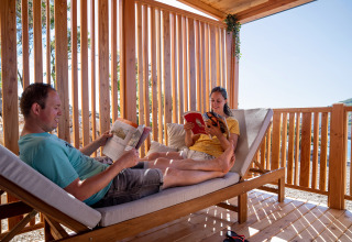 Dos personas se relajan y leen libros en una terraza soleada de madera en SunLodge BigLeaf, rodeada de naturaleza.