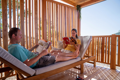 Dos personas se relajan y leen libros en una terraza soleada de madera en SunLodge BigLeaf, rodeada de naturaleza.