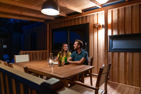Par nyder aften på træterrasse ved SunLodge BigLeaf, sidder ved oplyst bord med drikkevarer.