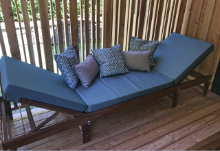 Udendørs lounge med blå hynder, træramme og puder, placeret på en terrasse med lodrette trælameller.