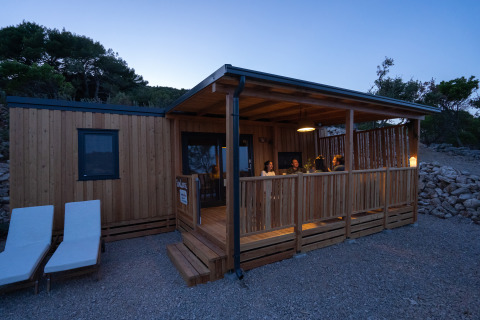 Aftenbillede af SunLodge BigLeaf med folk på terrassen og to liggestole foran hytten, omgivet af natur.