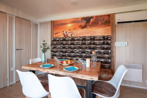 Comedor en SunLodge BigLeaf con mesa de madera, sillas blancas y decoración de pared africana al atardecer.