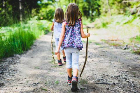 Two children walk on a forest path holding sticks during a glamping stay surrounded by greenery.