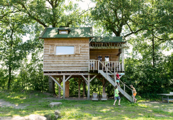 Children playing near a wooden treehouse on stilts, surrounded by trees, with stairs leading up.