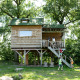 Kinder spielen bei einem Baumhaus auf Stelzen, umgeben von Bäumen, mit Zugangstreppe und Sonne.