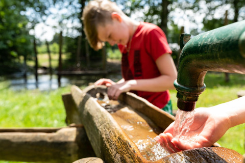 Ein Kind schöpft Wasser aus einem Außenhahn an einem Baumhaus im Beloofde Land, Niederlande, bei Sonnenschein.