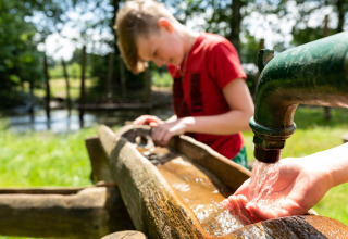 Ein Kind schöpft Wasser aus einem Außenhahn an einem Baumhaus im Beloofde Land, Niederlande, bei Sonnenschein.