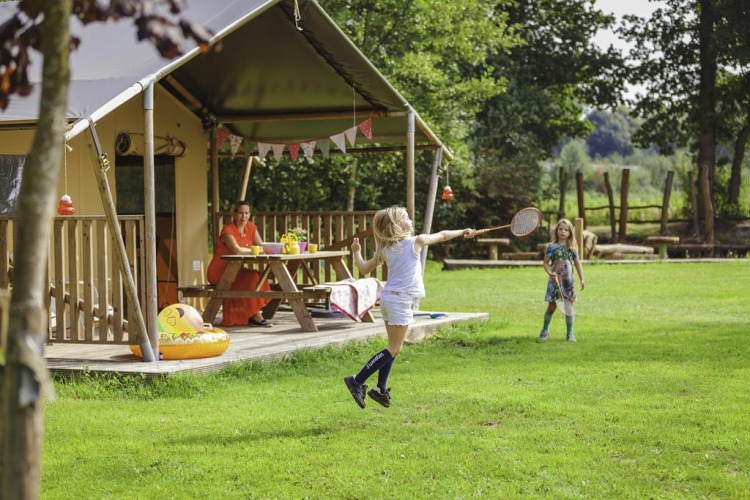 Kinderen spelen badminton voor een safaritent op Beloofde Land, Nederland, terwijl een vrouw toekijkt.
