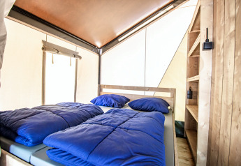 Sfeervol glamping-interieur met dubbel bed, blauwe dekens, houten rekken en veel lichtinval.