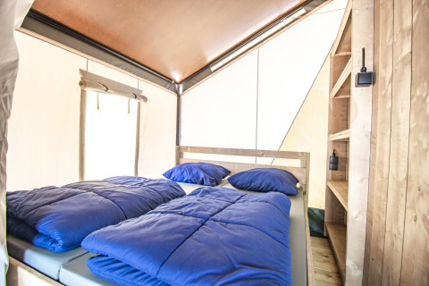 Interno di una tenda glamping con letto matrimoniale, biancheria blu e scaffali in legno chiaro.