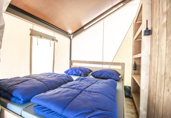Indretning af et glamping-telt med en dobbeltseng, blå sengetøj, træhylder og lyst interiør.