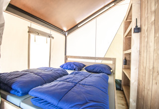 Interno di una tenda glamping con letto matrimoniale, biancheria blu e scaffali in legno chiaro.
