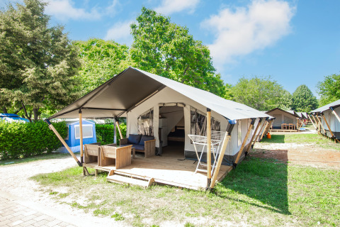 Tenda glamping moderna con veranda arredata, immersa nel verde sotto un cielo azzurro e soleggiato.