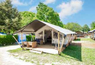 Tenda glamping moderna con veranda arredata, immersa nel verde sotto un cielo azzurro e soleggiato.