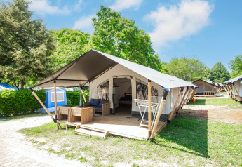 Modern glamping tent with wooden furniture on a deck, set in lush greenery under a bright blue sky.