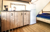 Indretning af et glamping-telt med trægulv, lille køkken, køleskab og dobbeltseng med blå sengetøj.