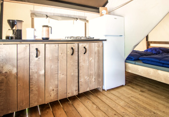 Binnenkant van een glamping tent met houten vloer, keukentje, koelkast en een bed met blauw dekbed.