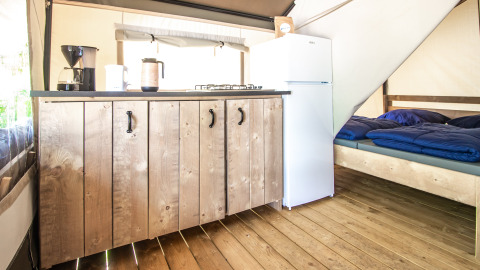 Interior de una tienda glamping con suelo de madera, cocina, nevera blanca y cama con mantas azules.