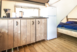 Interior de una tienda glamping con suelo de madera, cocina, nevera blanca y cama con mantas azules.