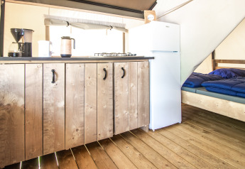 Interno di una tenda glamping con pavimento in legno, angolo cottura, frigo e letto con coperta blu.