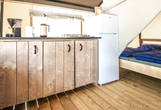 Interior de una tienda glamping con suelo de madera, cocina, nevera blanca y cama con mantas azules.
