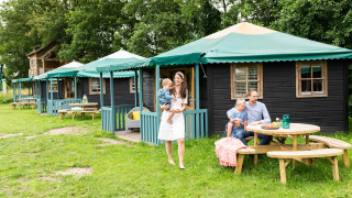 Familie nyder udendørs tid foran et naturhus kaldet Forest cottage med grønne tage og træborde.