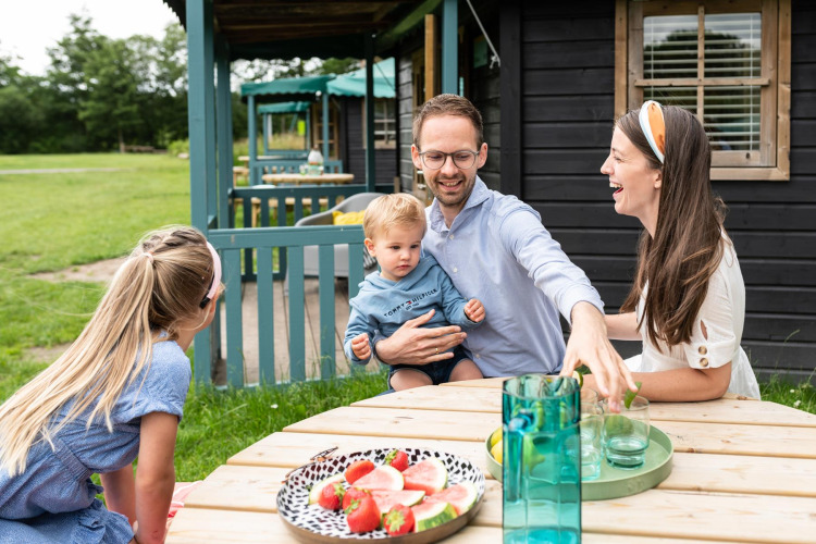 Familie hygger sig udenfor ved et bord med frugt ved skovhytte på Beloofde Land, Holland.