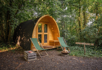 Casetta pod in legno immersa nel bosco con due sedie verdi, braciere e un cartello rustico tra gli alberi.