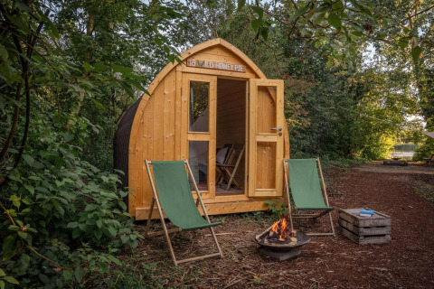 Eine Holzhütte mit offenen Türen, zwei grünen Liegestühlen und einer Feuerstelle im Wald.