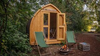 Cabaña de madera con puertas abiertas, dos tumbonas verdes y una hoguera en un entorno natural.