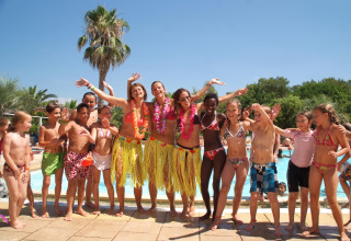 Children and adults in colorful swimwear and grass skirts celebrate by the pool at Camping Leï Suves in France.
