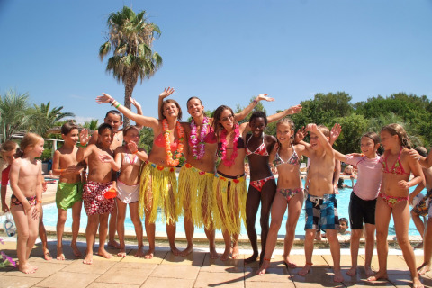 Children and adults in colorful swimwear and grass skirts celebrate by the pool at Camping Leï Suves in France.