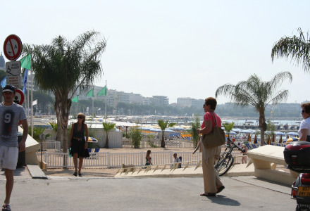 Paseo marítimo cerca de Roquebrune-sur-Argens con palmeras, gente caminando y vista costera al fondo.
