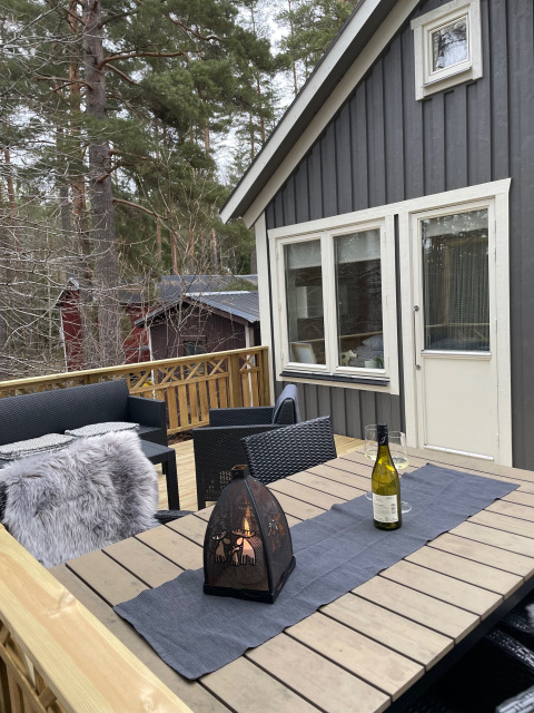 Terraza exterior en Småland Miniglamping, Suecia, con mesa, botella de vino, linterna y bosque de fondo.