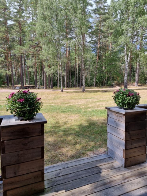 Vista desde una terraza de madera con macetas de flores hacia césped y bosque en Småland Miniglamping, Suecia.