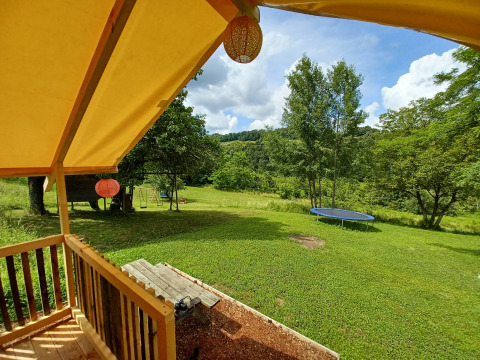 Blick vom Safari-Zelt auf eine grüne Wiese mit Trampolin, Schaukel und Bäumen unter blauem Himmel.