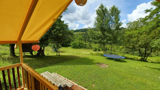 Blick vom Safari-Zelt auf eine grüne Wiese mit Trampolin, Schaukel und Bäumen unter blauem Himmel.