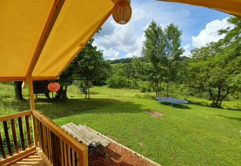 Blick vom Safari-Zelt auf eine grüne Wiese mit Trampolin, Schaukel und Bäumen unter blauem Himmel.
