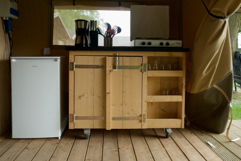 Compact kitchen setup in a safari tent with mini fridge, wooden cabinet, gas stove, and utensils on top.