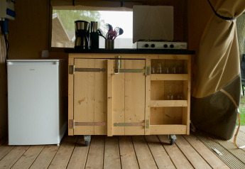 Pequeña cocina en una tienda safari con mini nevera, armario de madera, hornillo de gas y utensilios.