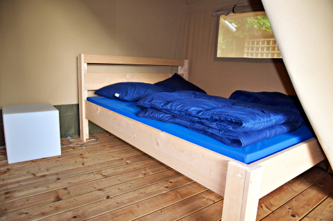 Interior de una tienda safari con una cama de madera, ropa de cama azul y una mesita de noche moderna.