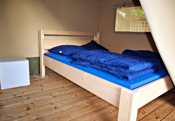 Intérieur d'une tente safari avec un lit en bois, literie bleue et une table de nuit cubique moderne.
