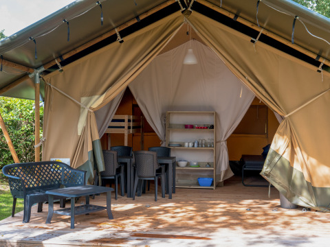Tienda safari con terraza cubierta, sillas y mesa de plástico, zona de dormir y comedor visibles en el interior.