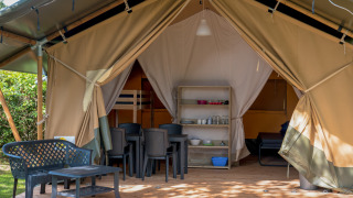 Tienda safari con terraza cubierta, sillas y mesa de plástico, zona de dormir y comedor visibles en el interior.