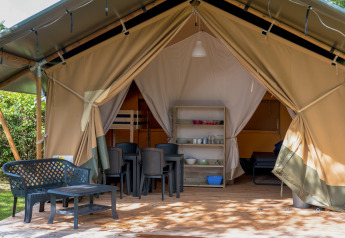 Tienda safari con terraza cubierta, sillas y mesa de plástico, zona de dormir y comedor visibles en el interior.