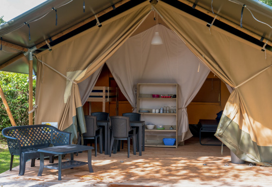 Tienda safari con terraza cubierta, sillas y mesa de plástico, zona de dormir y comedor visibles en el interior.