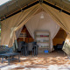 Tienda safari con terraza cubierta, sillas y mesa de plástico, zona de dormir y comedor visibles en el interior.