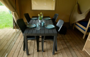 Dining table inside a safari tent, set for four with plates and glasses, plastic chairs and wooden floor.