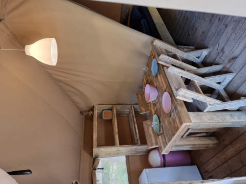 Interno di una tenda safari con mobili rustici in legno, tavolo da pranzo, sedie, piatti e lampada.